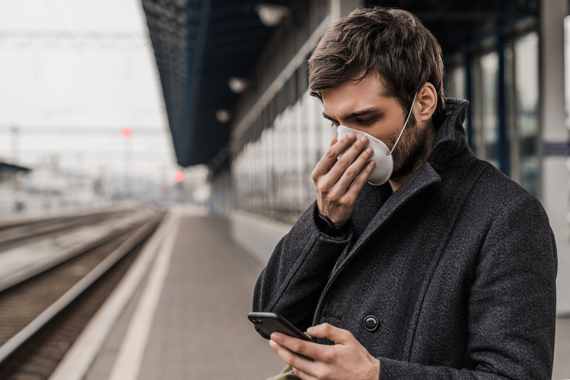 Mann mit Maske steht am Bahnsteig und blickt auf sein Handy.